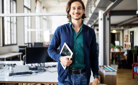 Junger lächelnder Mann in offenem Büro mit einem Tablet in der Hand.