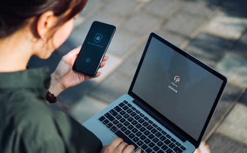 Eine sitzende Frau mit Laptop auf dem Schoß und Smartphone in der linken Hand, die über ihr Smartphone eine Sicherheitsauthentifzierung durchführt.