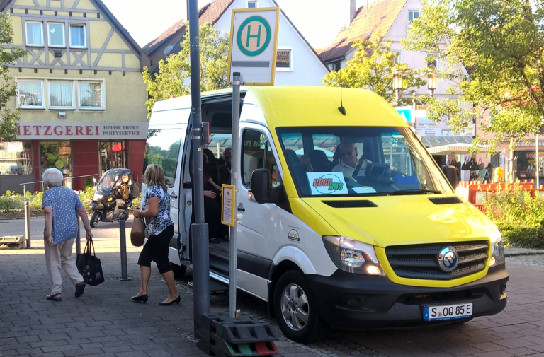 Gelb-weißer Kleinbus steht an Bushaltestelle in einem Dorf.