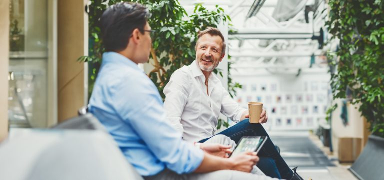 Zwei Männer sitzen auf einer Bank und unterhalten sich. Der eine hält einen Pappbecher, der andere einen Tablet-Computer.