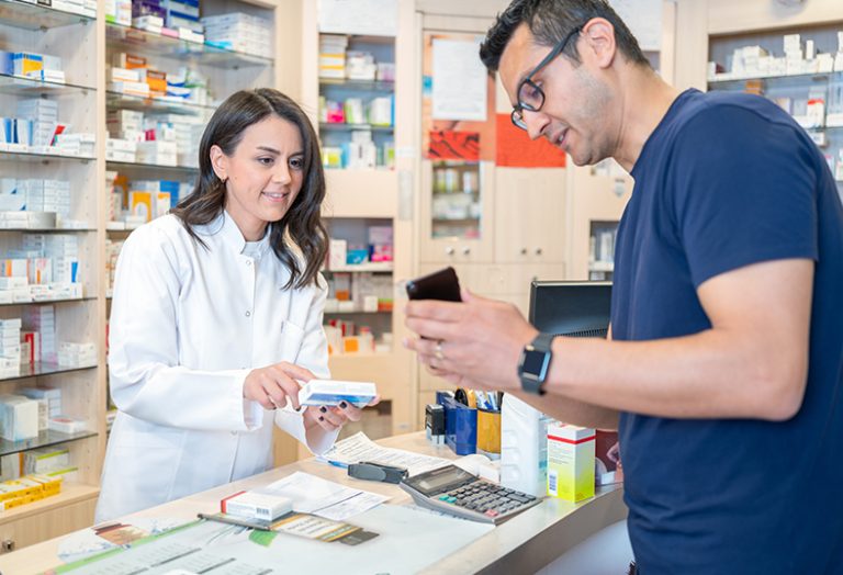 Mann zeigt Frau in weißem Kittel in einer Apotheke sein Smartphone.