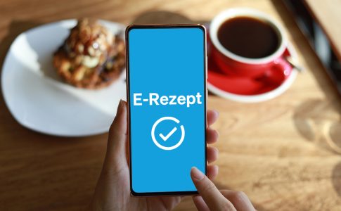 Smartphone mit symbolischem e-Rezept Schriftzug in blau auf dem Bildschirm vor Holztisch mit Kuchenstück und Kaffeetasse