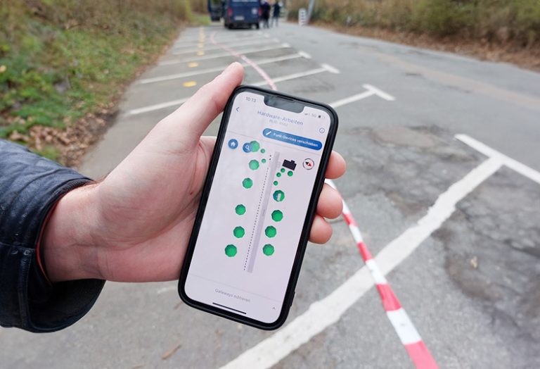 Ein Smartphone-Bildschirm zeigt in einer App die Belegung eines Parkplatzes an. Im Hintergrund sind die Parkplätze mit den gelben Sensoren zu sehen.