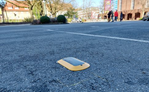 Ein grauer Sensor mit gelbem Rahmen ist auf dem Asphalt befestigt. Er meldet Daten zur Parkplatzbelegung.