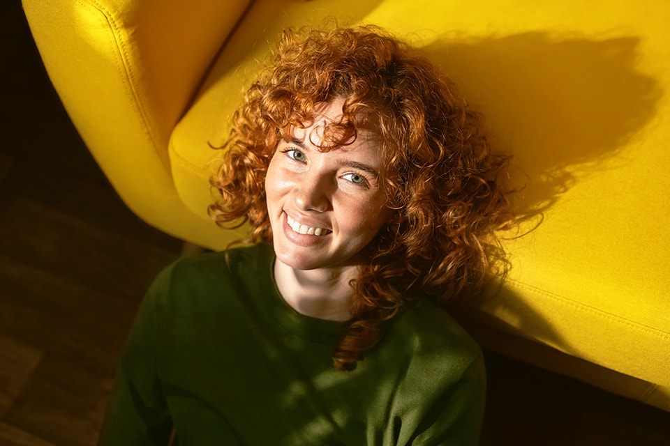 Lächelnde Frau mit roten Locken und grünem Pullover sitzt vor einem gelben Sessel auf dem Boden.